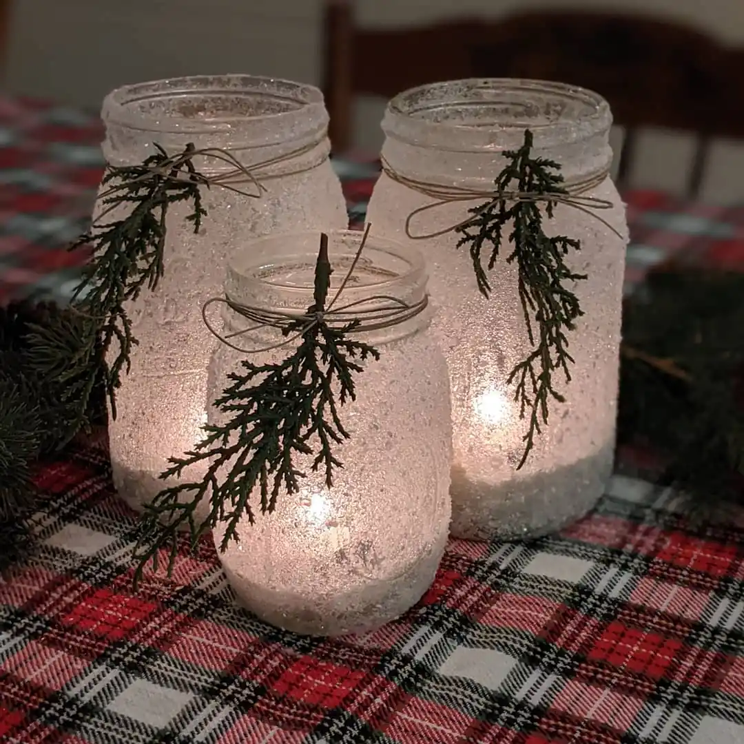 Three frosted mason jars glowing with candlelight and decorated with small evergreen sprigs and twine