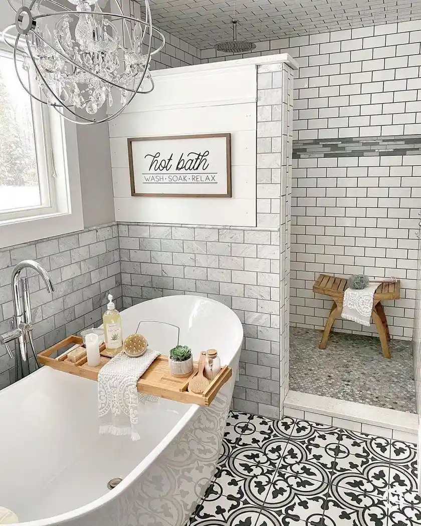 Modern farmhouse bathroom with freestanding tub, patterned black-and-white floor tile, marble-look walls, wood bath tray, and crystal chandelier