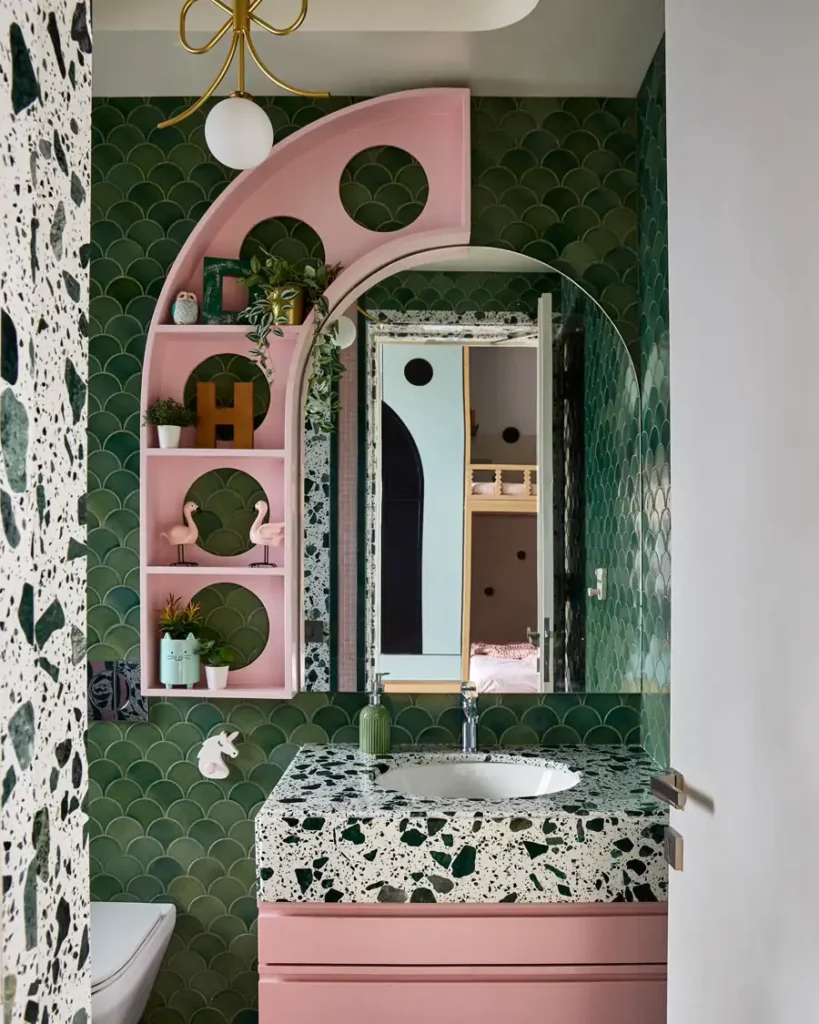 Playful bathroom with green scallop tiles, curved pink shelving, a large mirror, terrazzo sink, and pink vanity.