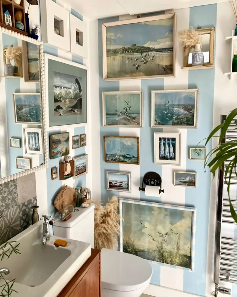 Coastal bathroom gallery wall with seascapes, shorebird paintings, and vintage ocean artwork on blue and white striped walls.