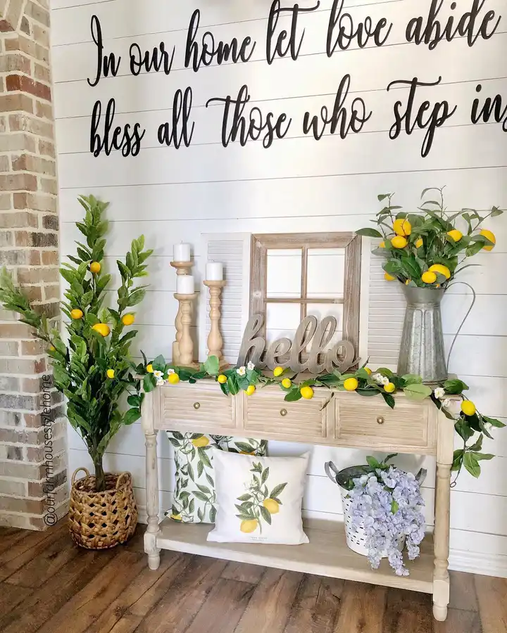 Light wood farmhouse entryway console decorated with lemon branches, greenery, tall candleholders, metal pitcher, botanical accents, and yellow details.