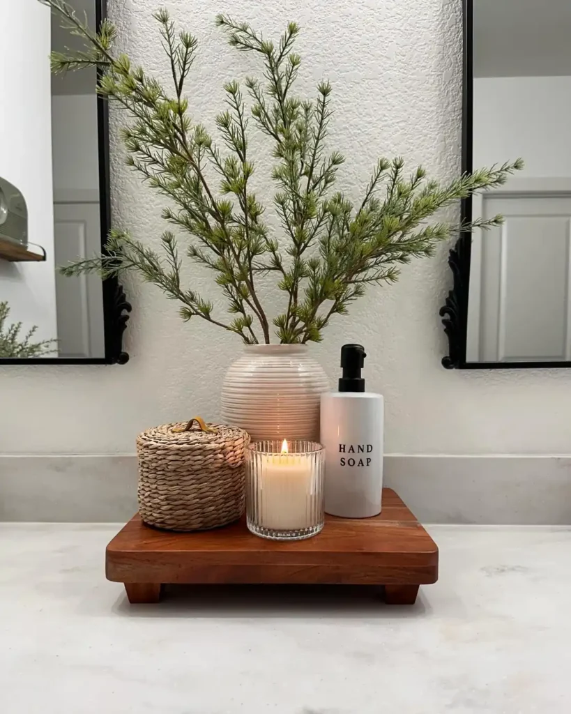 Bathroom counter decor on a small wooden riser with leafy branches, woven lidded container, candle, and white soap dispenser.
