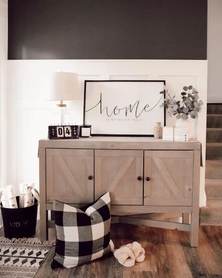 Light wood entryway cabinet with framed home artwork, lamp, candle, leafy stems, plaid pillow, slippers, and practical storage.