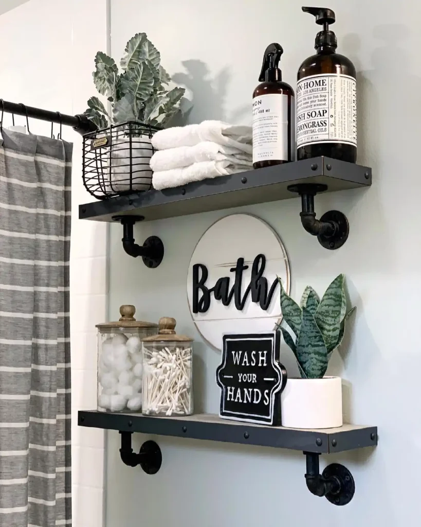 Industrial bathroom shelves with black pipe brackets, folded towels, amber bottles, cotton jars, plants, and wall signs.