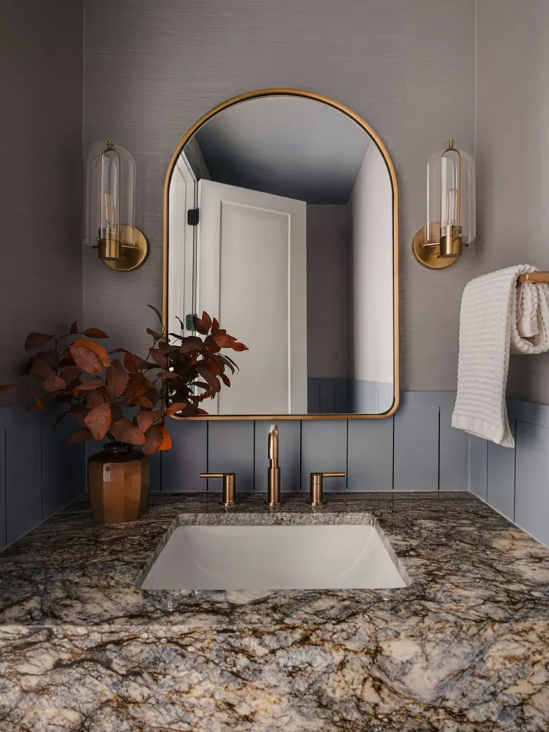 Moody bathroom vanity with dark stone counter, bronze fixtures, and a single vase filled with rust colored leafy branches.