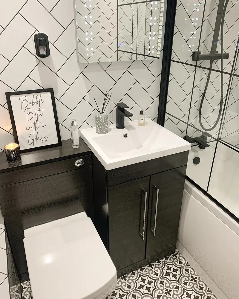 Compact black-and-white bathroom with herringbone wall tiles, a dark vanity, patterned floor, and grid shower screen.