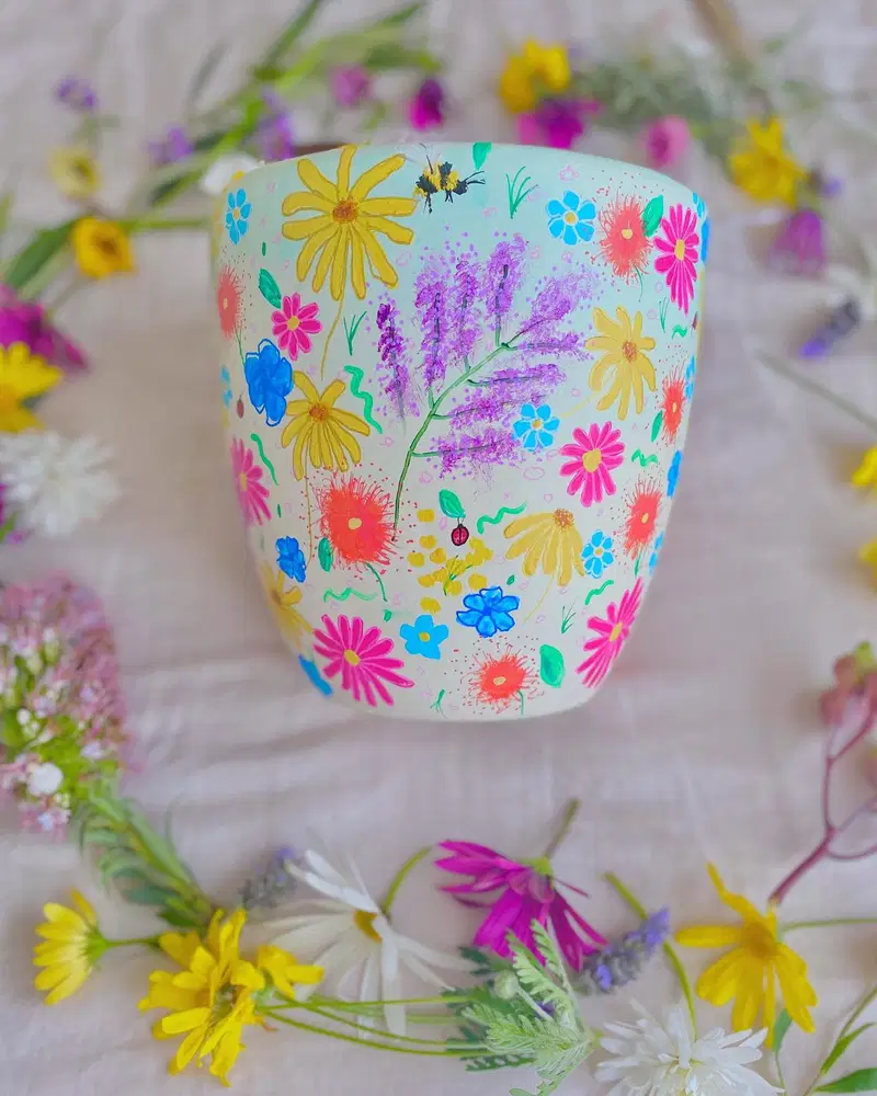 Light flower pot covered with colorful wildflowers, lavender sprigs, tiny leaves, and painted insects