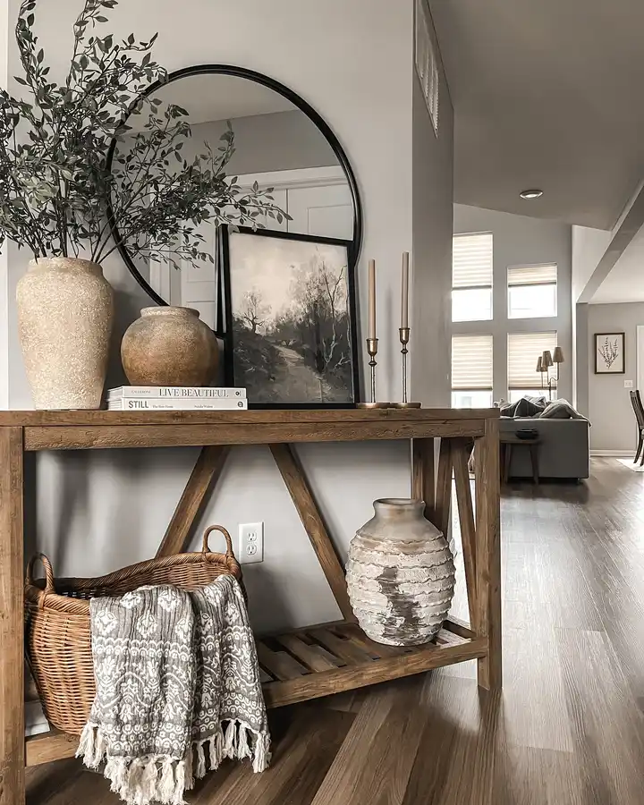 Rustic entryway console with a round black mirror, oversized neutral pottery, leafy branches, candlesticks, woven basket, and patterned throw.