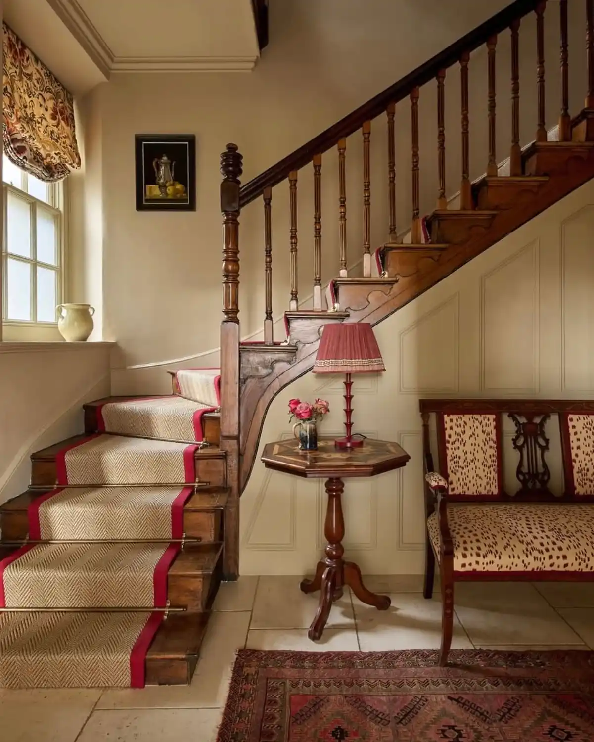 Traditional stair hallway with red trimmed stair runner, patterned rug, pleated lamp shade, and warm wood details