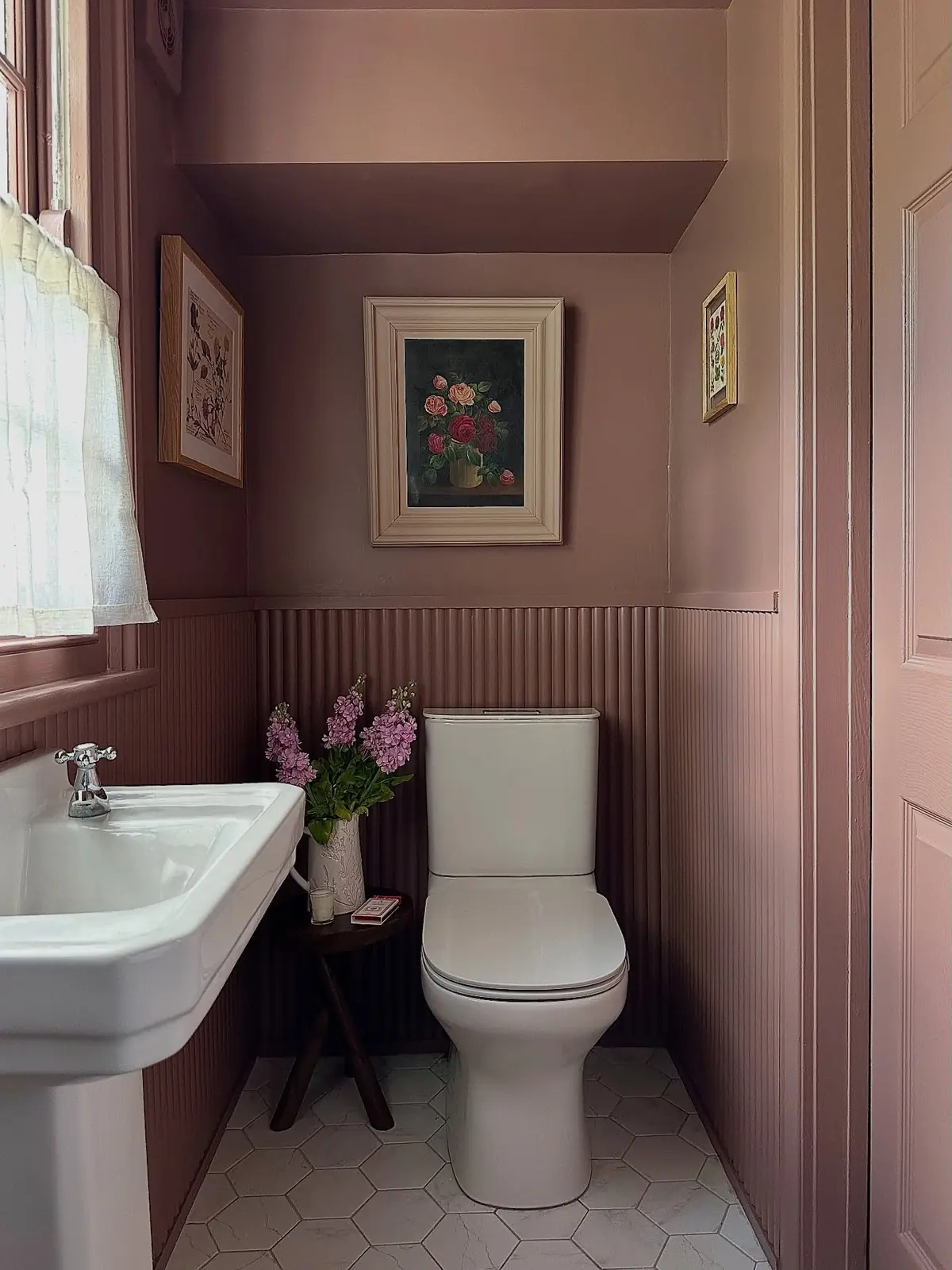 Narrow dusty pink bathroom with matching trim, ribbed paneling, a pedestal sink, floral artwork, and a window.