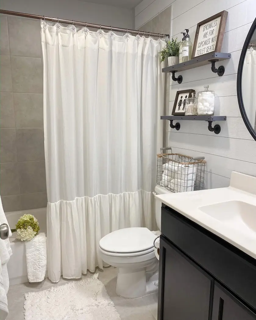 Small modern farmhouse bathroom with black vanity, white ruffled shower curtain, pipe shelves, shiplap wall, glass jars, and wire basket
