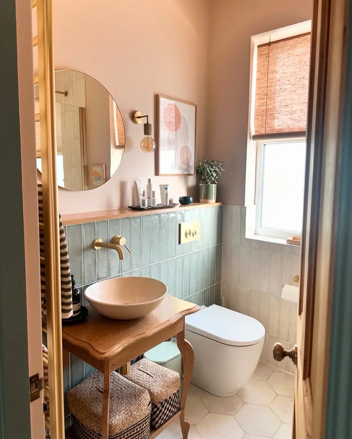Small pastel bathroom with an open-leg wooden vanity, bowl sink, woven storage baskets, and a round mirror.