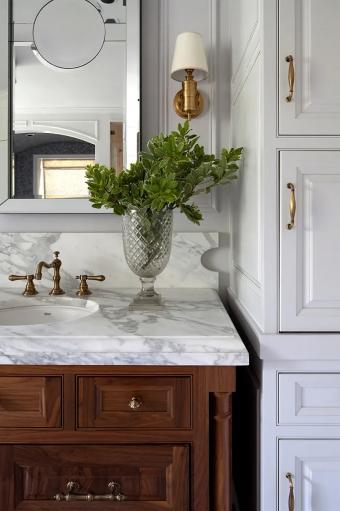 Marble bathroom vanity with dark wood cabinetry, brass faucet, and a tall cut glass vase filled with fresh green branches.