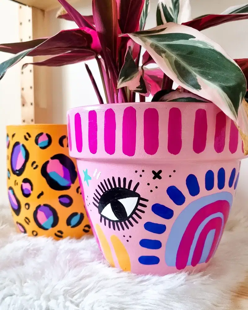 Pink flower pot painted with a graphic eye, rainbow arches, colorful brush marks, and playful symbols