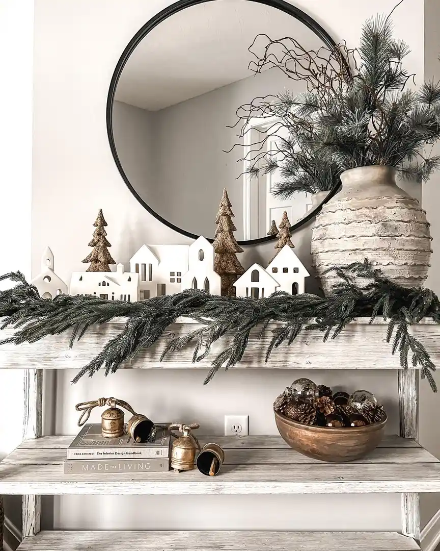 Winter entryway console with small white village houses, bronze trees, evergreen garland, oversized textured vase, black round mirror, books, bells, and pinecones.