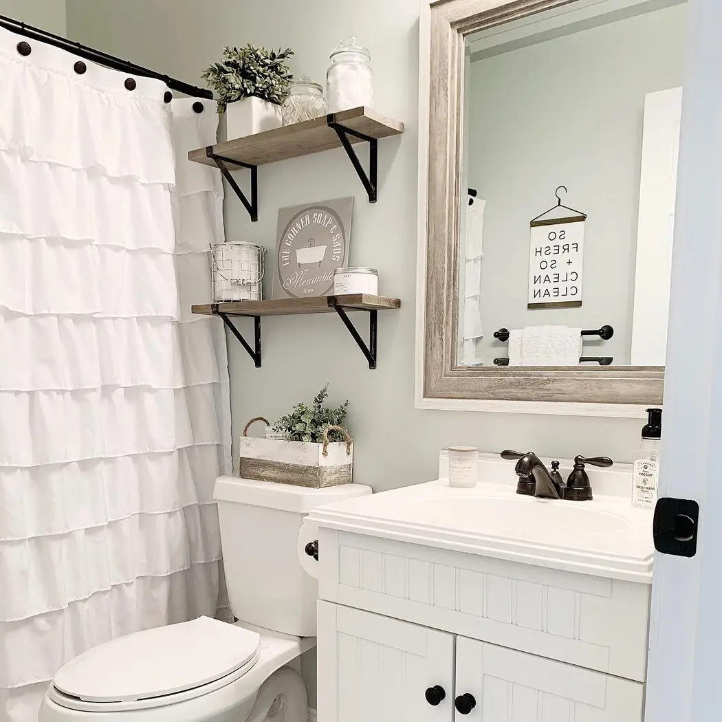 Cozy farmhouse bathroom with white ruffled shower curtain, rustic wood shelves, black brackets, pale green-gray walls, and weathered mirror