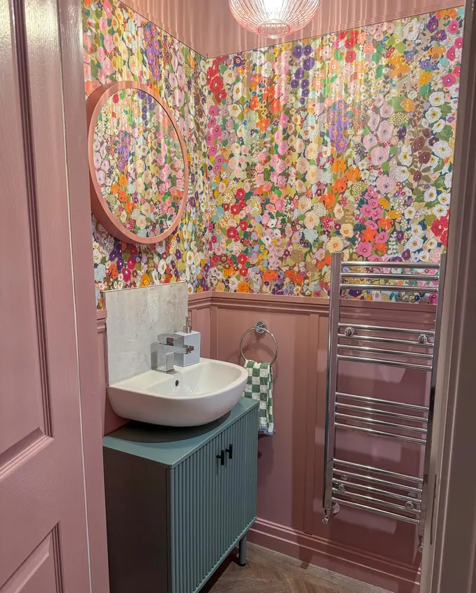 Tiny colorful powder room with floral wallpaper, pink paneling and ceiling, a teal vanity, and a round mirror.