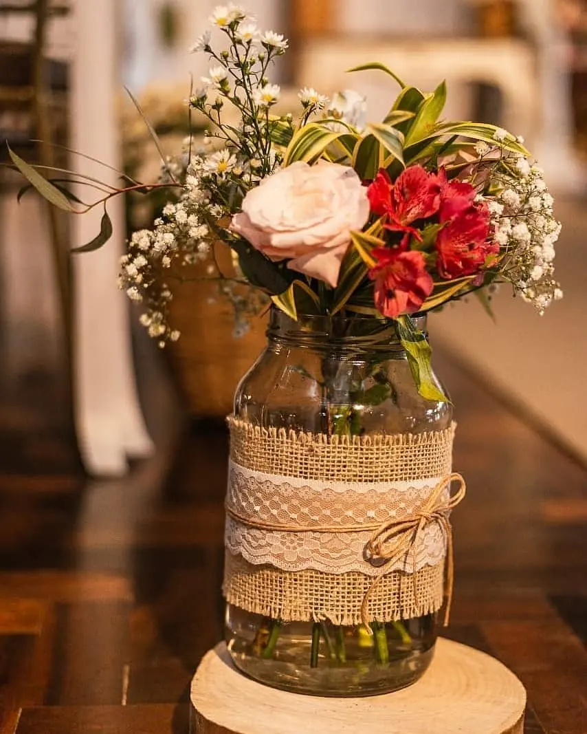 Mixed flowers and greenery in a mason jar wrapped with burlap, lace, and twine on a wood slice