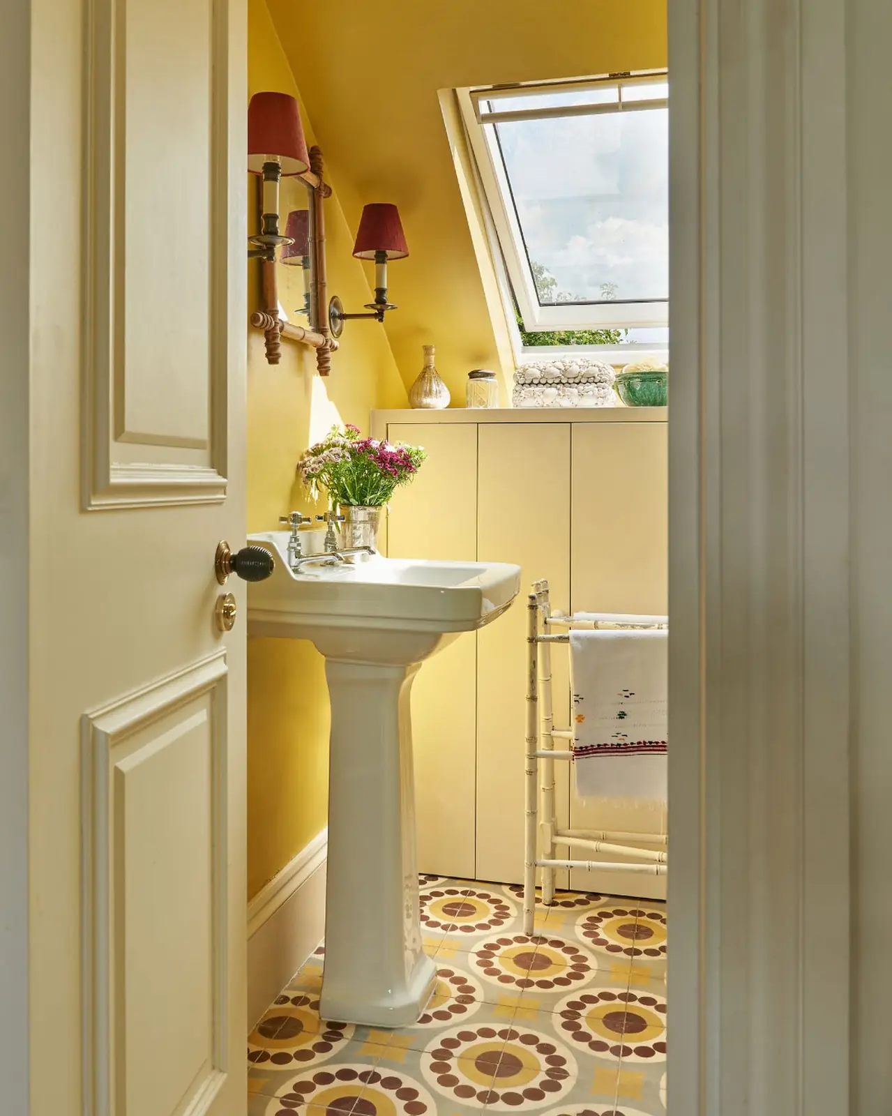 Tiny yellow attic bathroom with a skylight, pedestal sink, fitted storage cabinet, and patterned floor tiles.