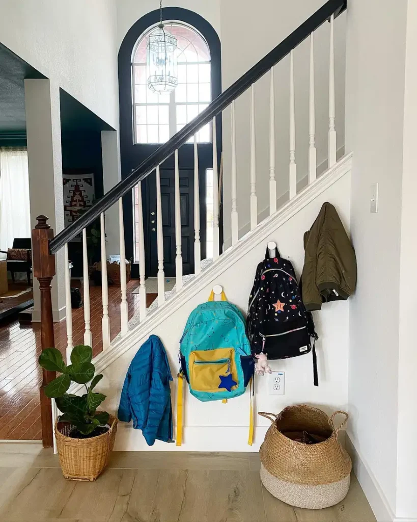 Backpacks and jackets hanging on hooks beneath a staircase with a woven basket for extra entryway gear.