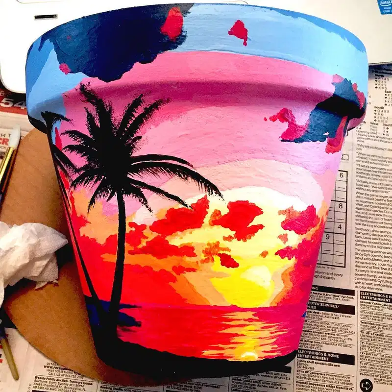 Flower pot painted with a colorful tropical sunset, glowing water, and black palm tree silhouette