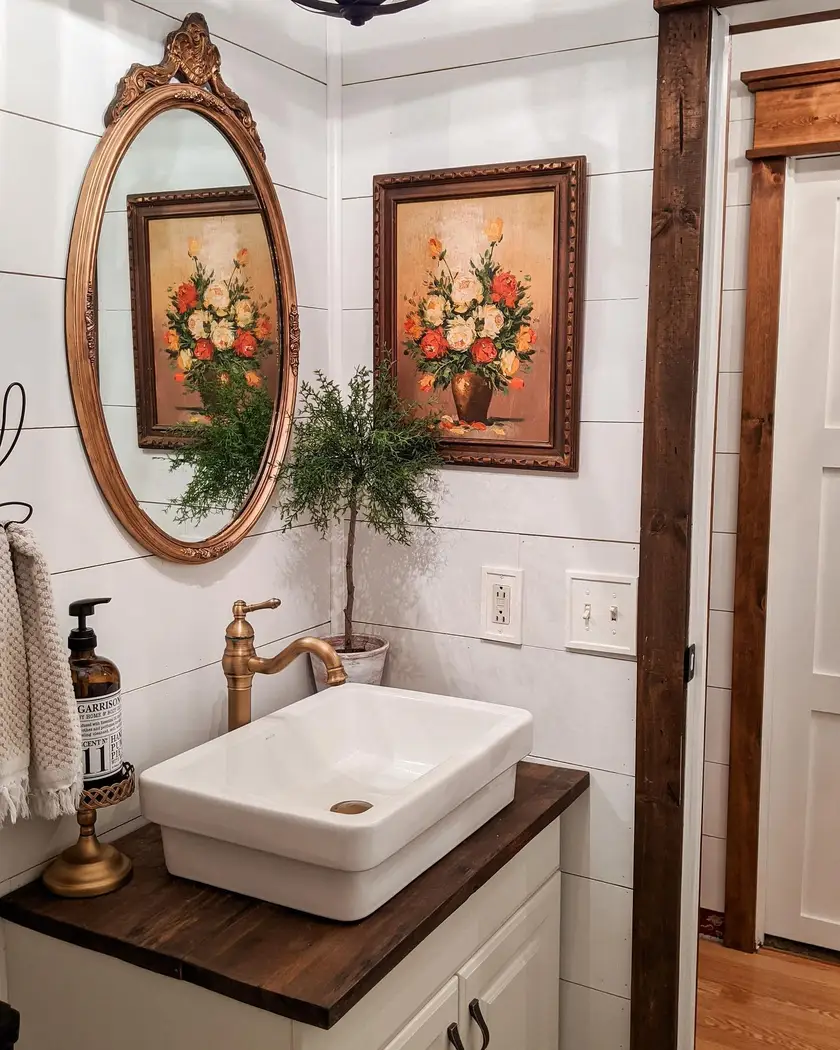 Vintage farmhouse bathroom vanity with dark wood countertop, white vessel sink, ornate oval mirror, floral artwork, and aged brass faucet