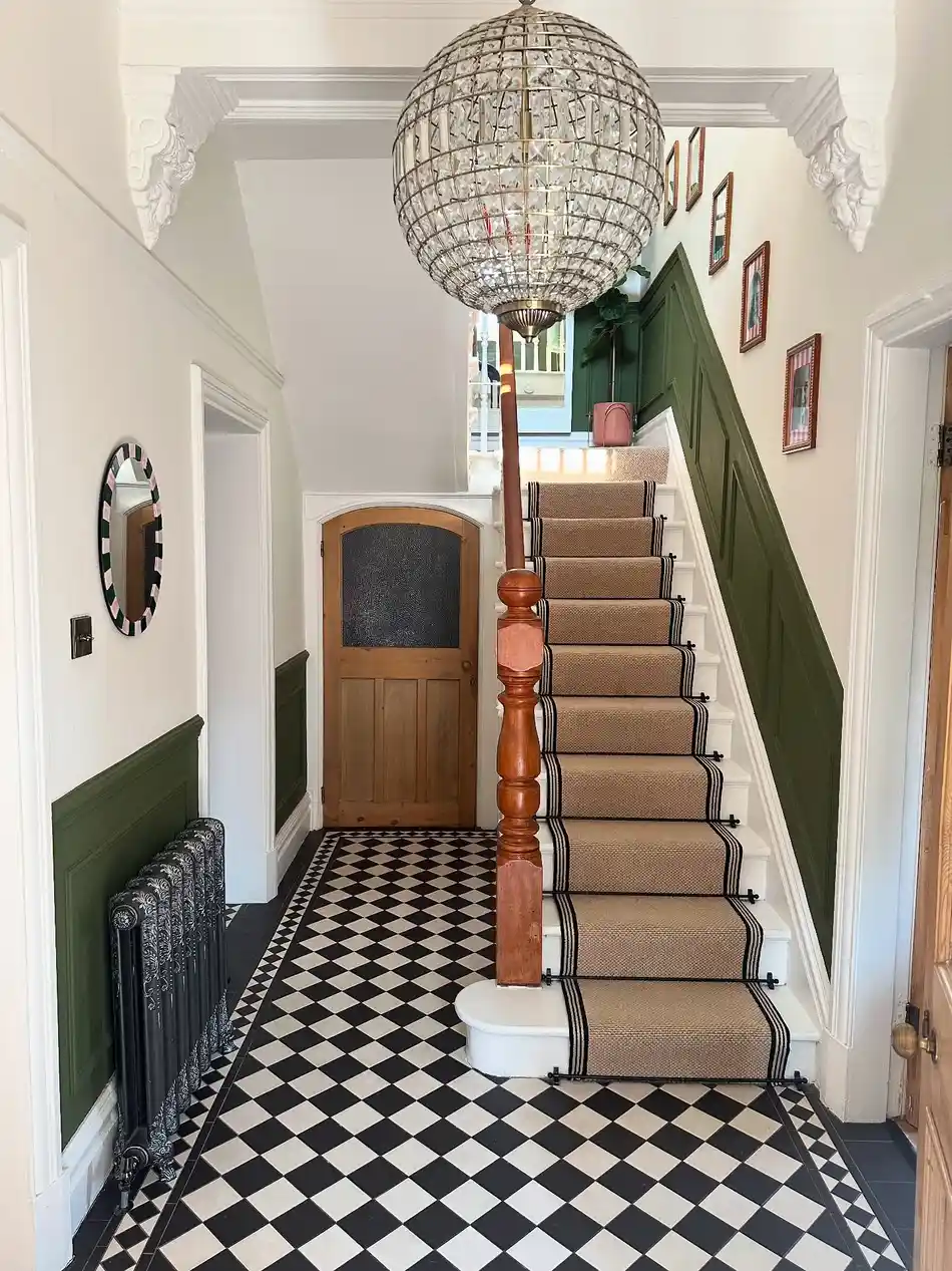Hallway with black and white checkerboard floor, deep green paneling, wood staircase, globe pendant, and neutral runner