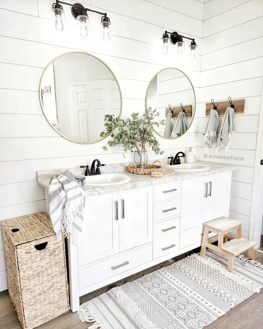 Bright modern farmhouse double vanity with brass-framed round mirrors, white shiplap, woven laundry basket, layered rugs, and black faucets