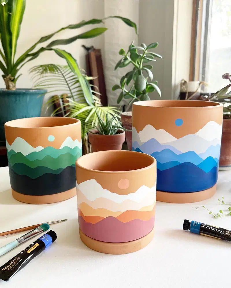 Terracotta flower pots painted with layered mountain landscapes in green, blue, and sunset tones