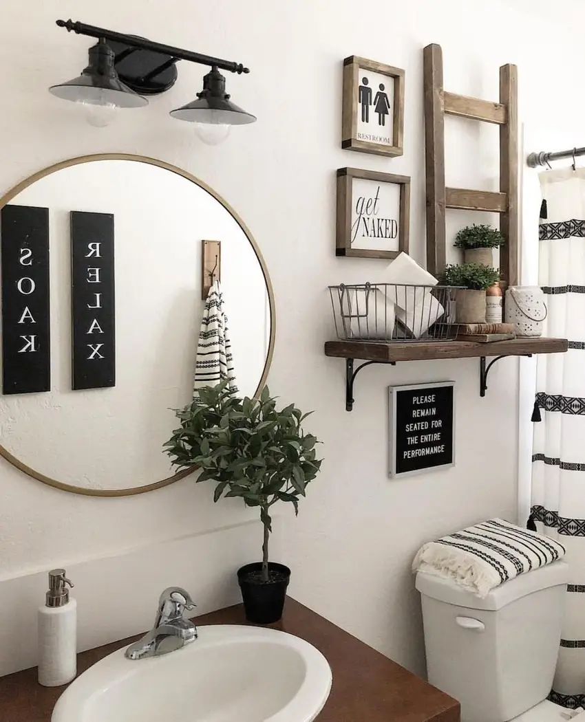 Modern farmhouse bathroom corner with rustic wood ladder, floating shelf, wire basket, framed wall art, round mirror, and greenery