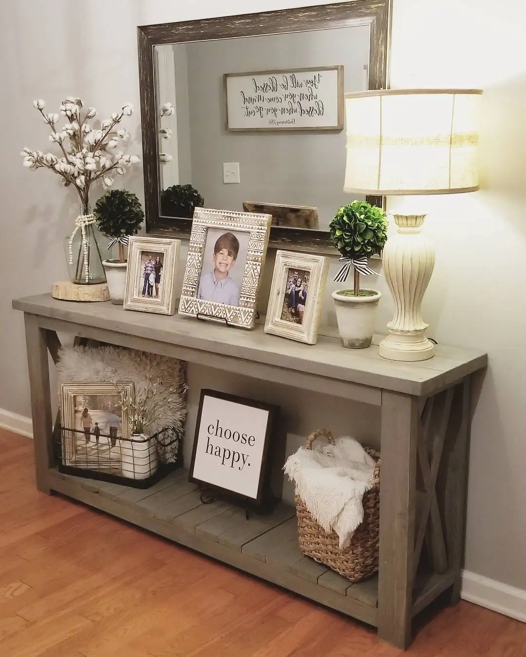 Hallway console with rustic mirror, grouped family photos, table lamp, cotton stems, small plants, basket, and folded throw