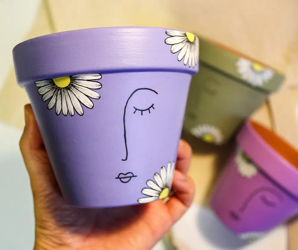 Lavender flower pot decorated with a minimalist line drawn face and white daisies around the edges