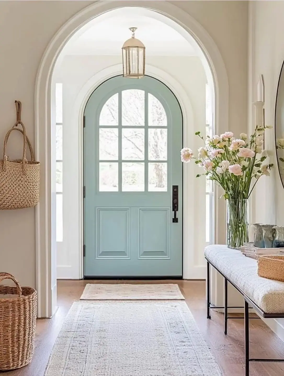 Soft neutral entry hallway with arched pale blue door, upholstered bench, woven baskets, runner, and pastel flowers