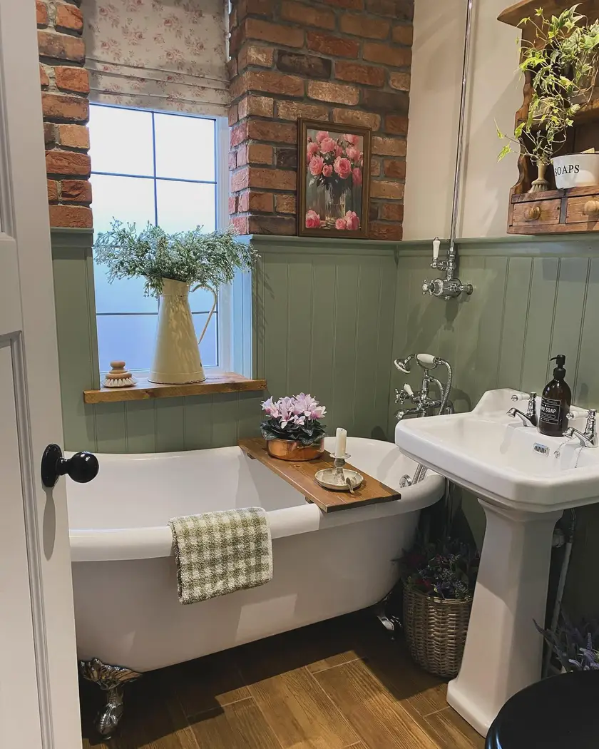 Cozy farmhouse bathroom with sage green paneling, exposed brick wall, clawfoot tub, pedestal sink, and warm wood accents