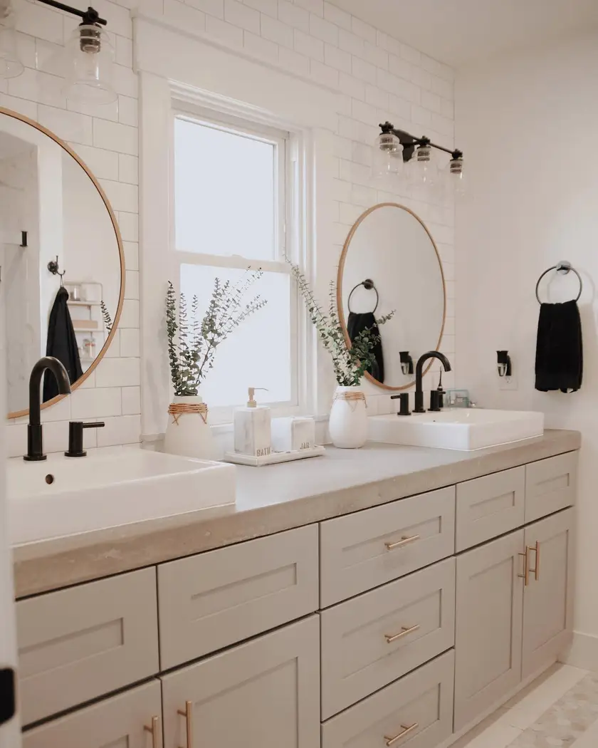 Bright modern farmhouse double vanity with white cabinets, black faucets, brass hardware, round mirrors, and simple greenery