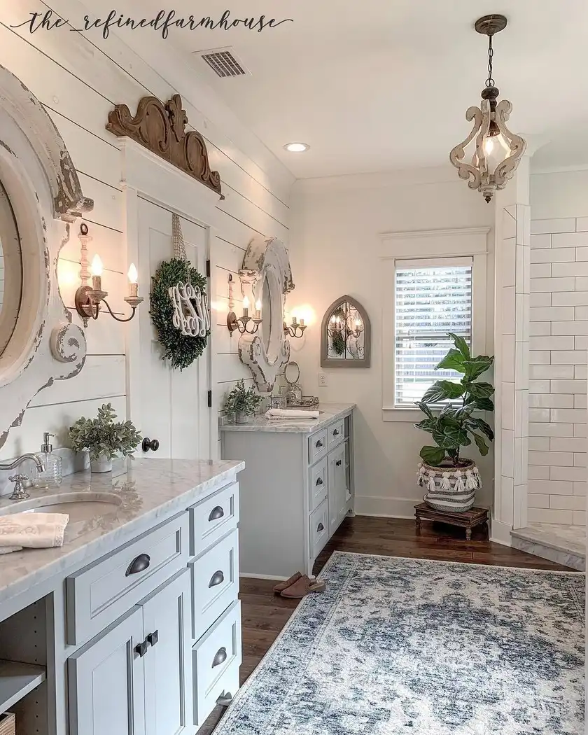 Elegant farmhouse bathroom with distressed mirrors, white shiplap, blue-gray vanities, vintage wall lights, chandelier, and patterned rug