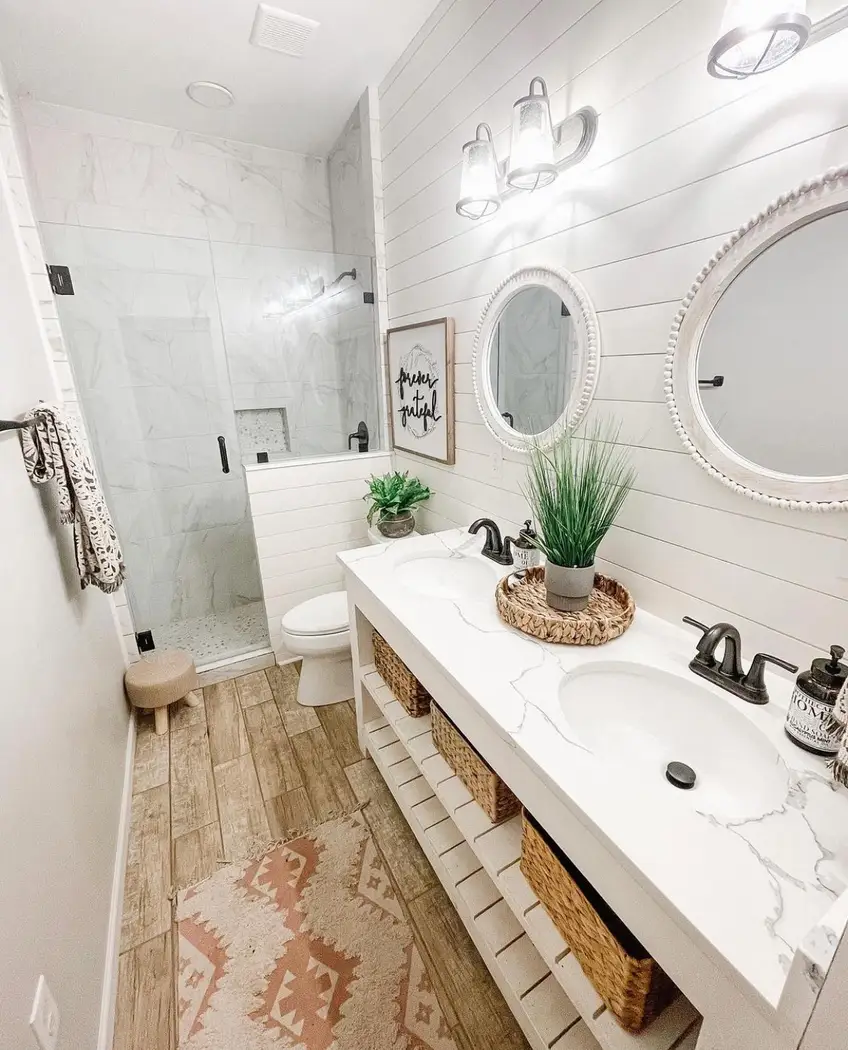 Modern farmhouse bathroom with white double vanity, open shelf storage, woven baskets, round mirrors, black faucets, and wood-look flooring