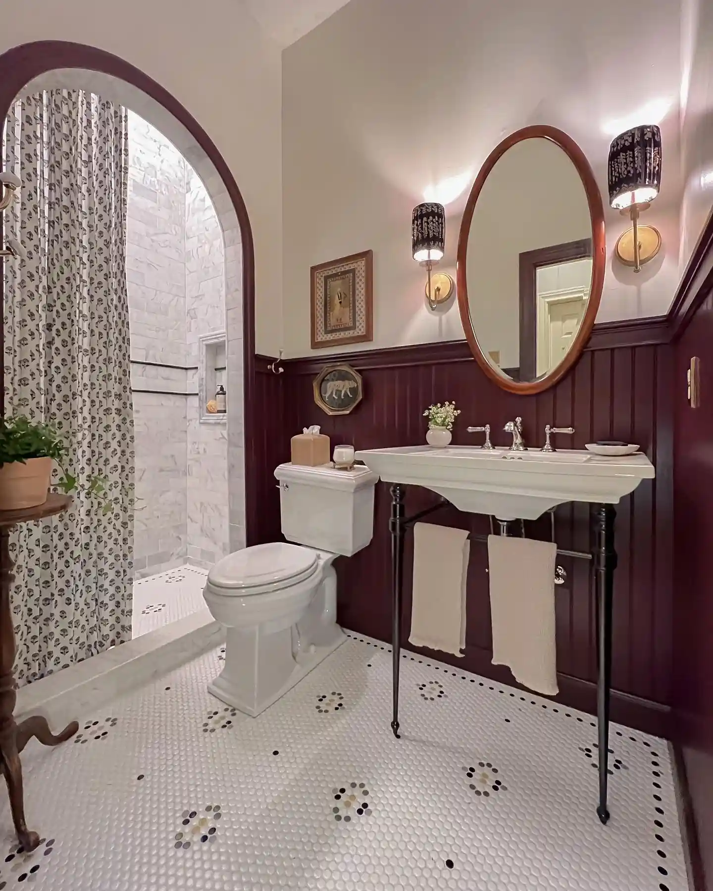 Vintage guest bathroom with deep burgundy wall paneling, oval mirror, white console sink, arched shower and penny tile floor