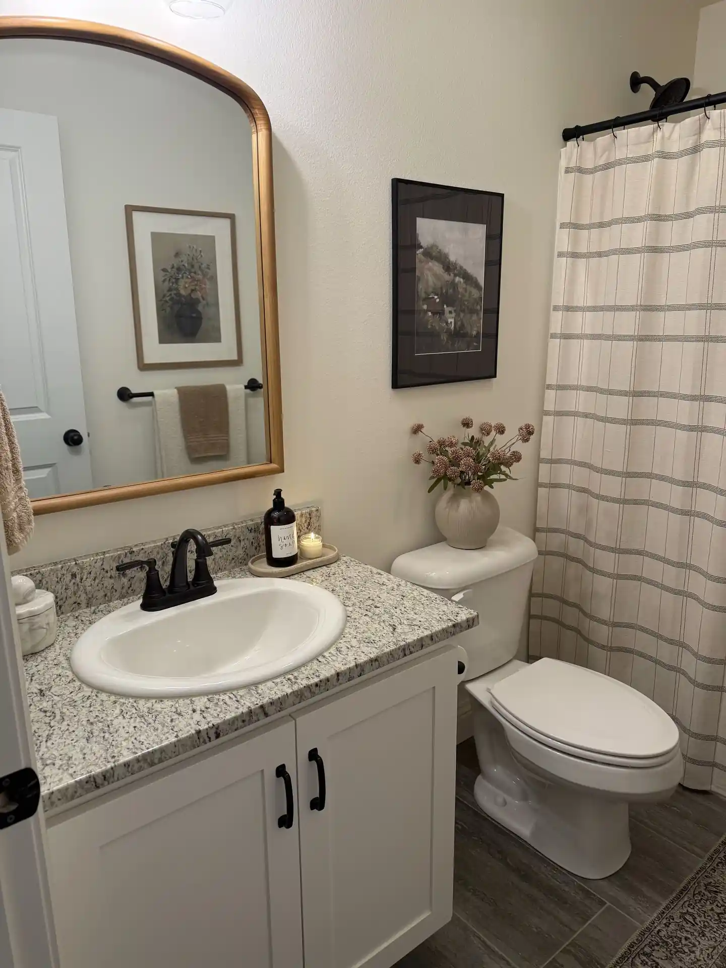 Simple guest bathroom with wood-framed mirror, white vanity, black fixtures, striped shower curtain, framed art and small flower vase