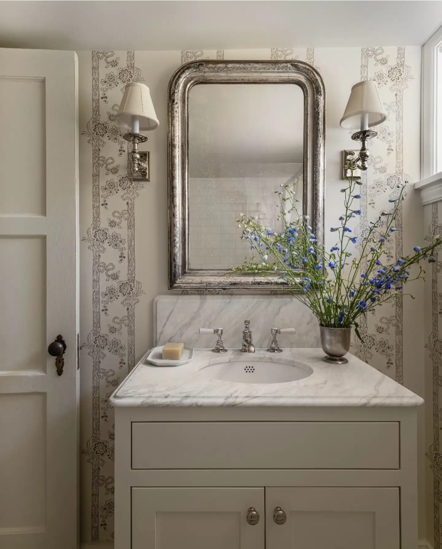 Soft neutral guest bathroom with floral stripe wallpaper, aged silver mirror, marble vanity, matching sconces and blue flowers