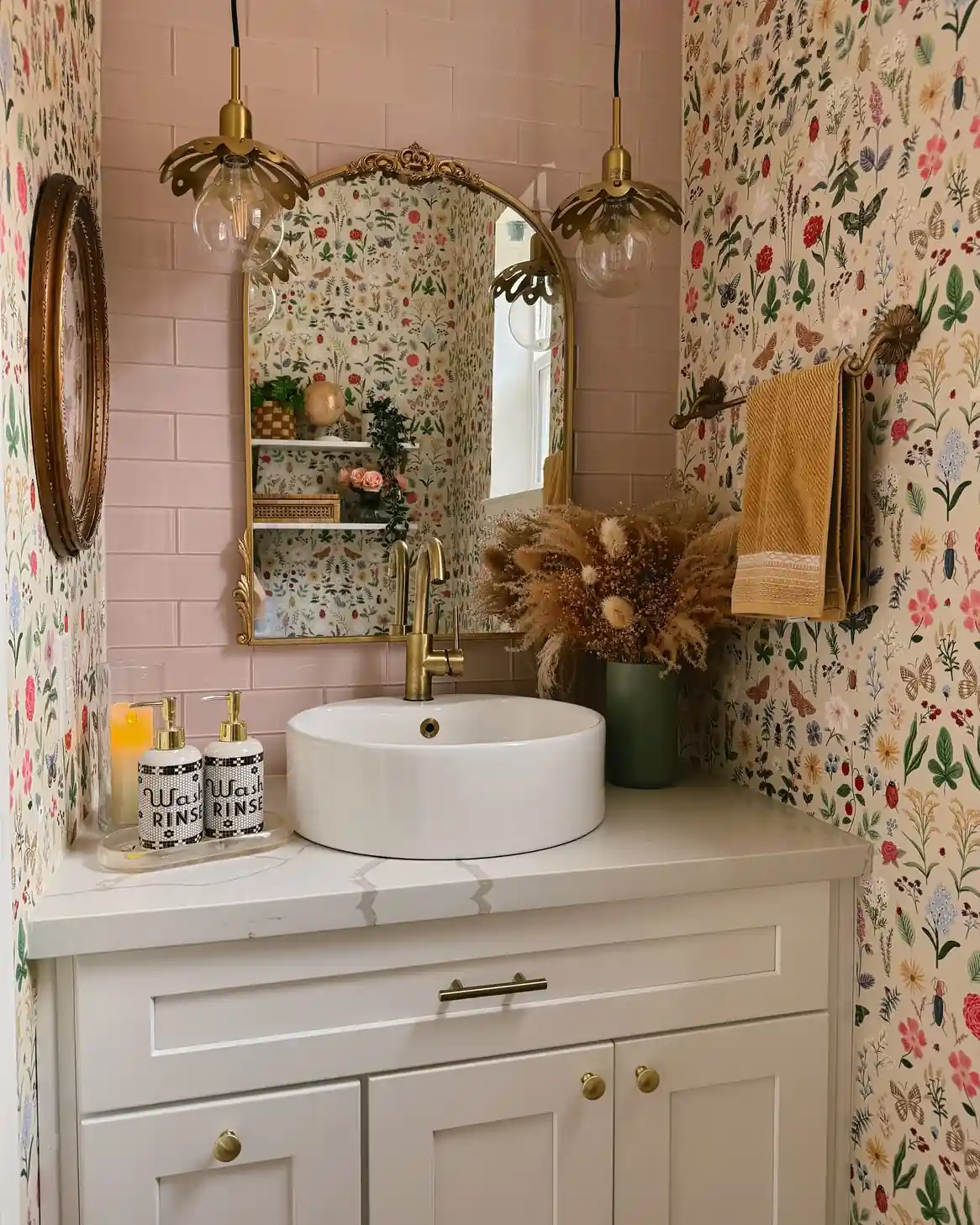 Colorful guest bathroom with botanical wallpaper, blush tile, white vanity, brass fixtures, pendant lights and mustard hand towel