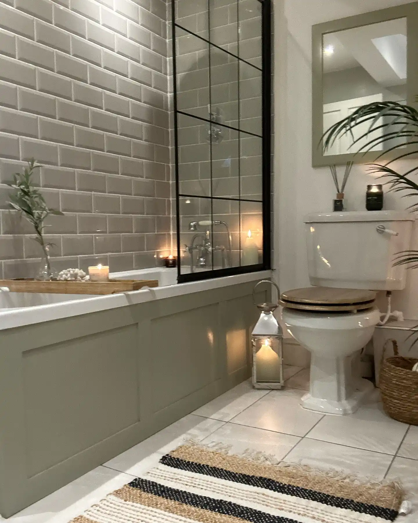 Cozy guest bathroom with sage green paneling, white subway tile, black shower screen, woven basket, striped rug and candles