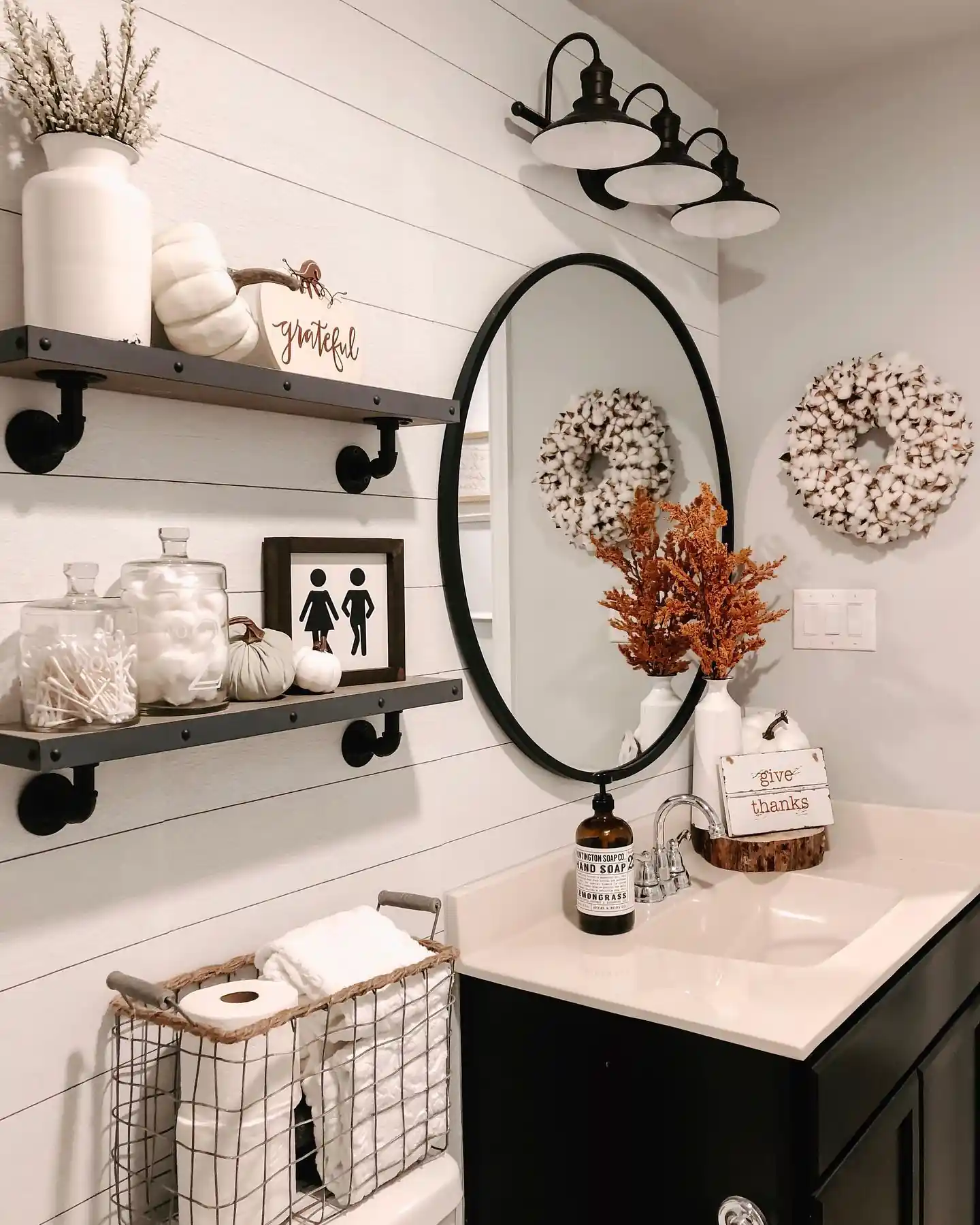 Farmhouse guest bathroom with white shiplap, black shelves, oval mirror, fall pumpkins, cotton wreaths and wire towel basket