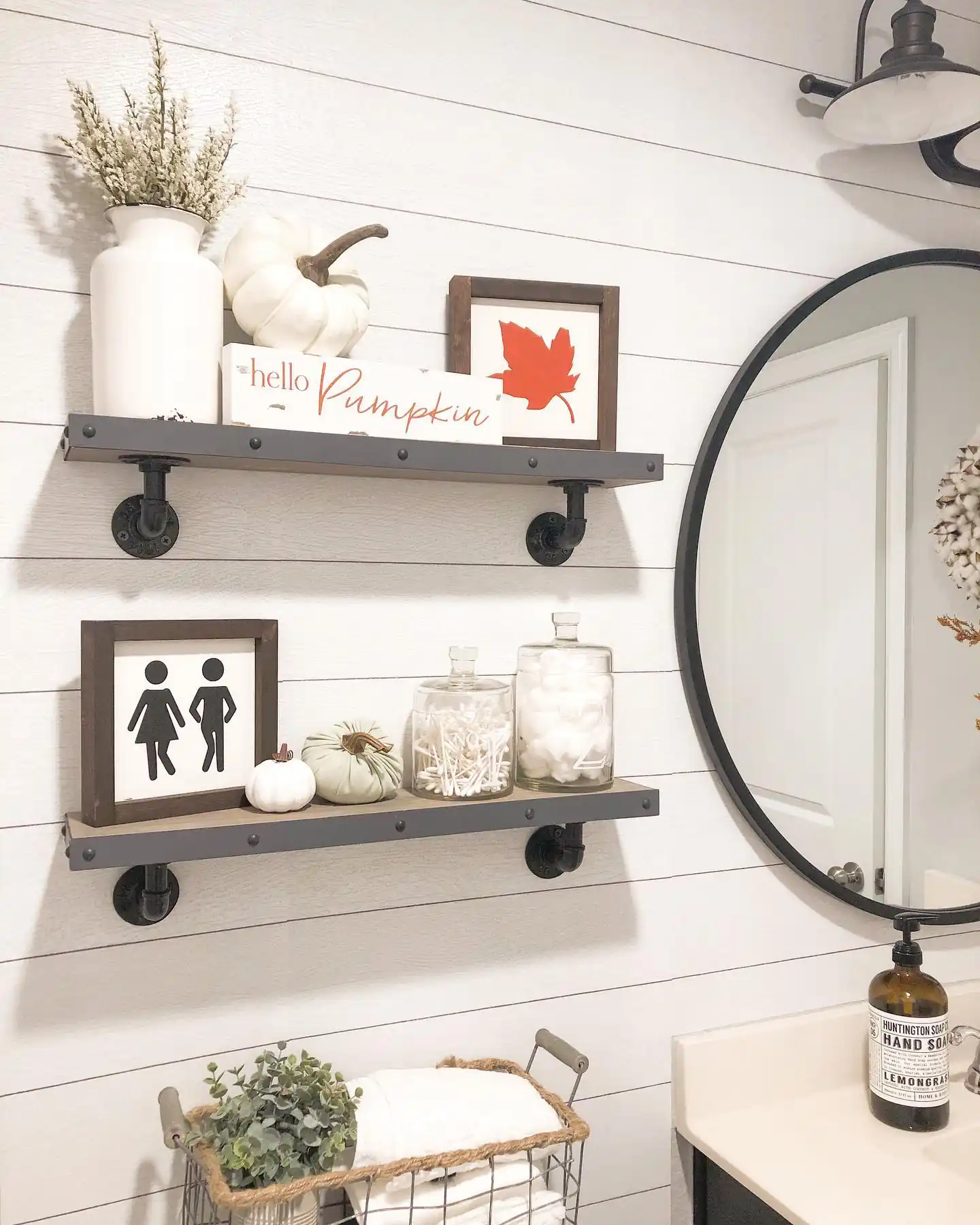 Farmhouse guest bathroom with white shiplap, black floating shelves, fall pumpkins, cotton jars, round mirror and folded towels
