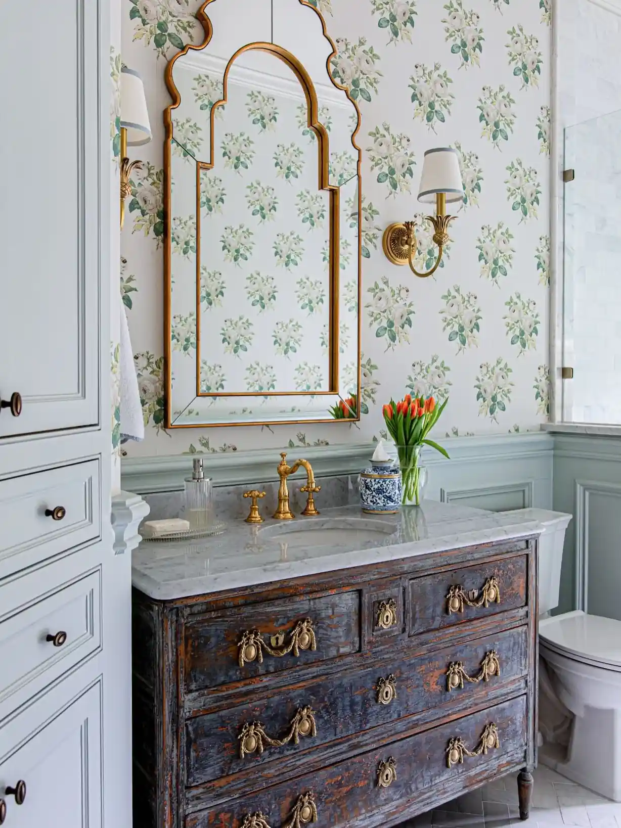 Guest bathroom with distressed vintage dresser vanity, floral wallpaper, marble countertop, brass fixtures and ornate gold mirror