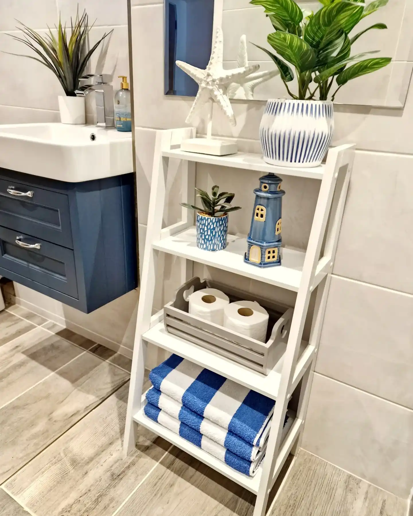 Guest bathroom with white ladder shelf holding blue towels, spare toilet paper, plants and coastal decor beside navy vanity
