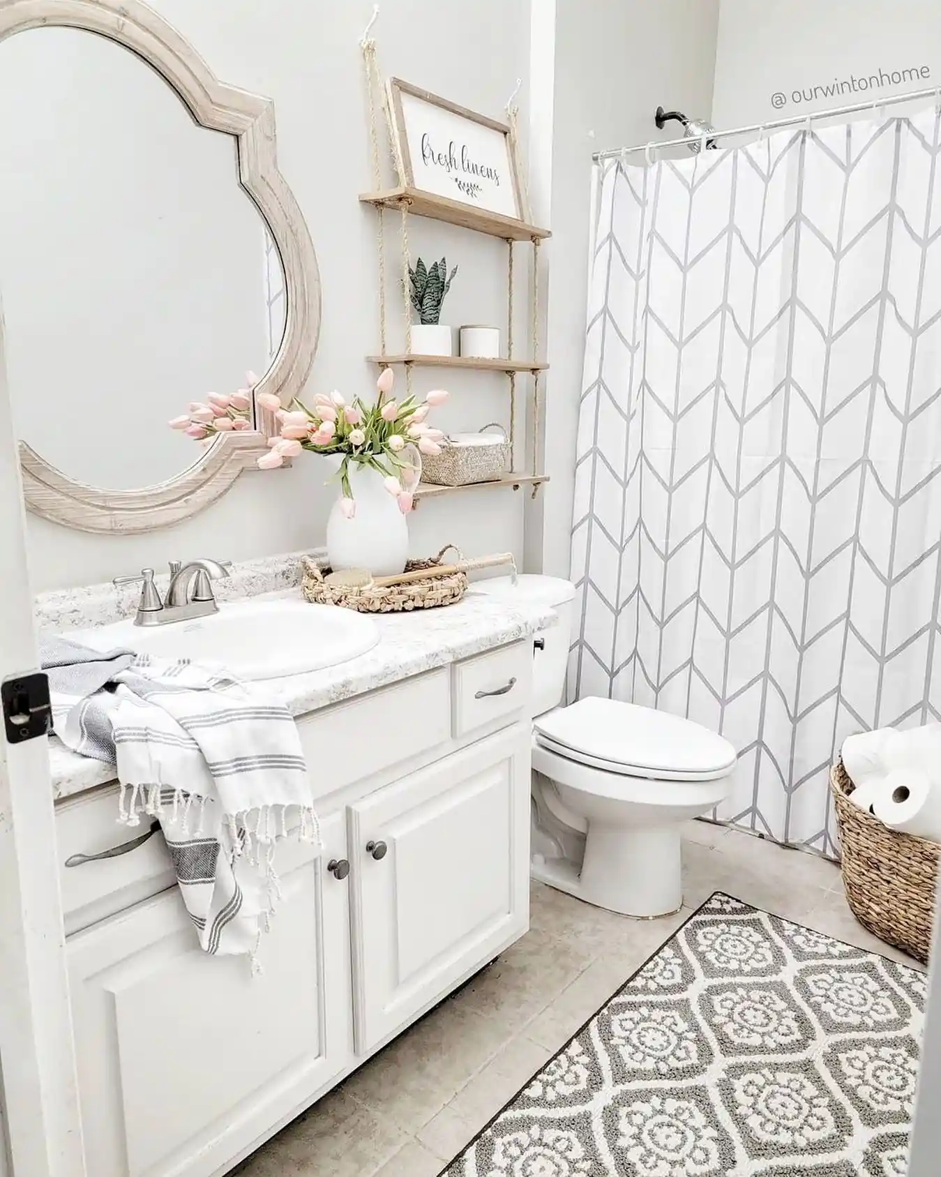 Bright white guest bathroom with wood-framed mirror, rope shelves, woven baskets, pink tulips, striped towel and patterned rug
