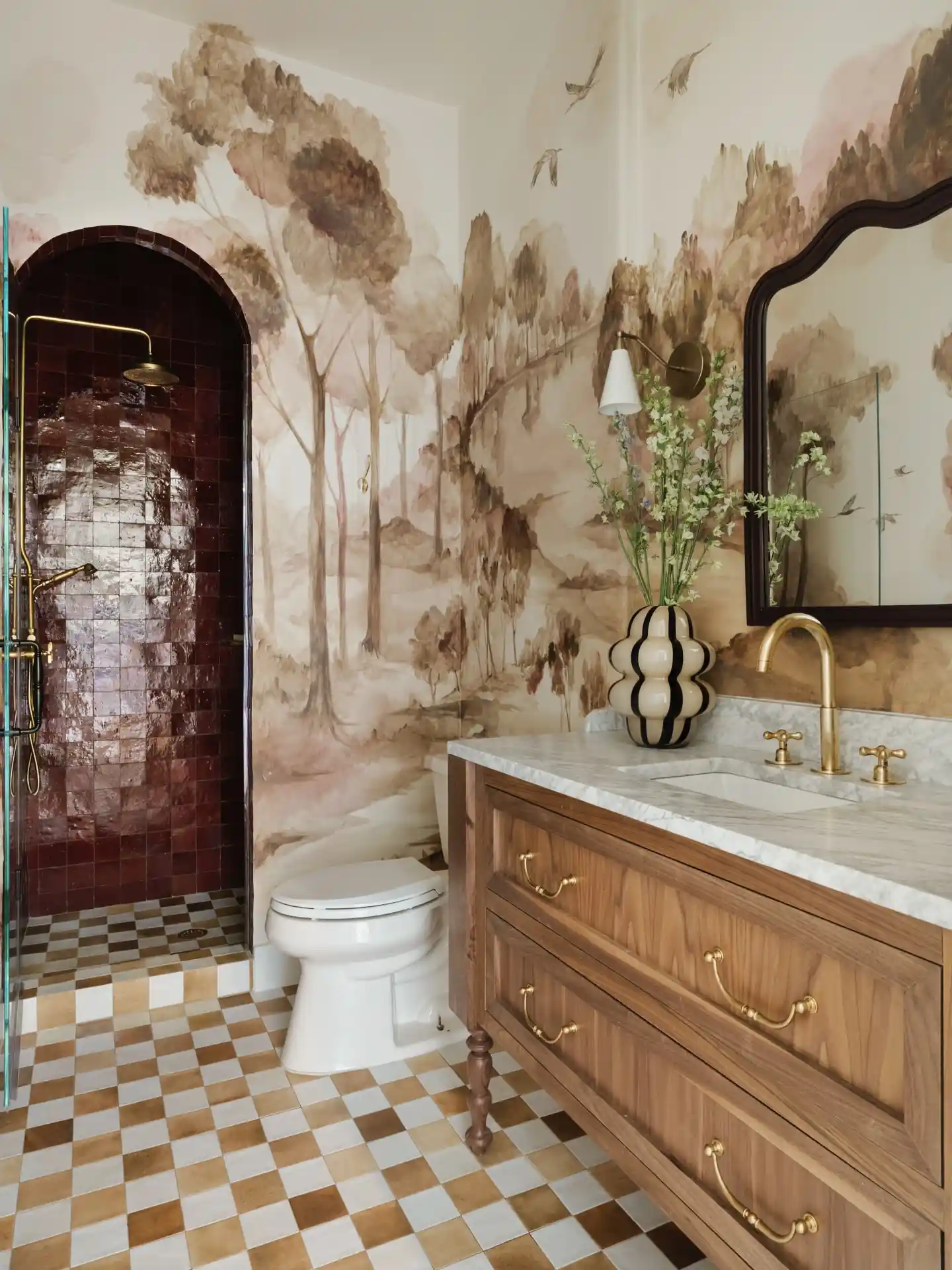 Guest bathroom with painterly landscape mural, burgundy tiled shower, checkerboard floor, wood vanity and brass fixtures