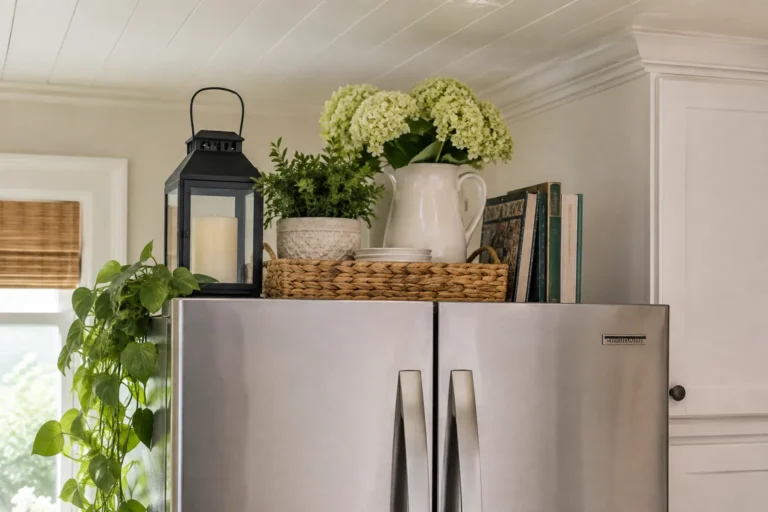 Stainless steel refrigerator topped with a woven tray, white flowers, potted greenery, cookbooks, and a black lantern in a bright farmhouse-style kitchen.
