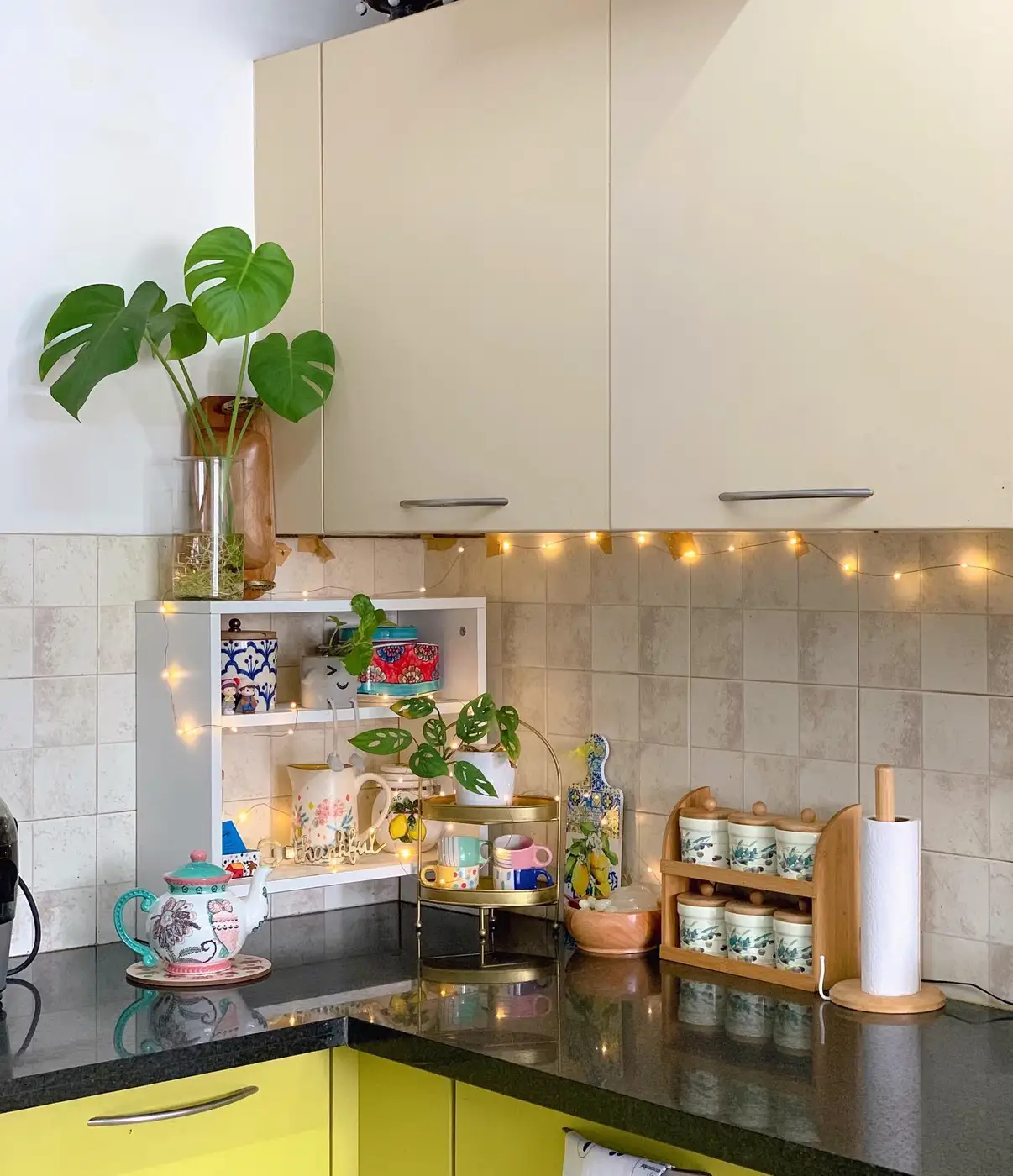 Tall green monstera stems, colorful mugs, teapot and warm string lights decorating a kitchen counter corner